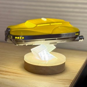 May include: A yellow futuristic vehicle model with a dark grey trim, mounted on a light-up stand. The stand has a wooden base and a clear, angular support. The vehicle has a sleek, aerodynamic design.
