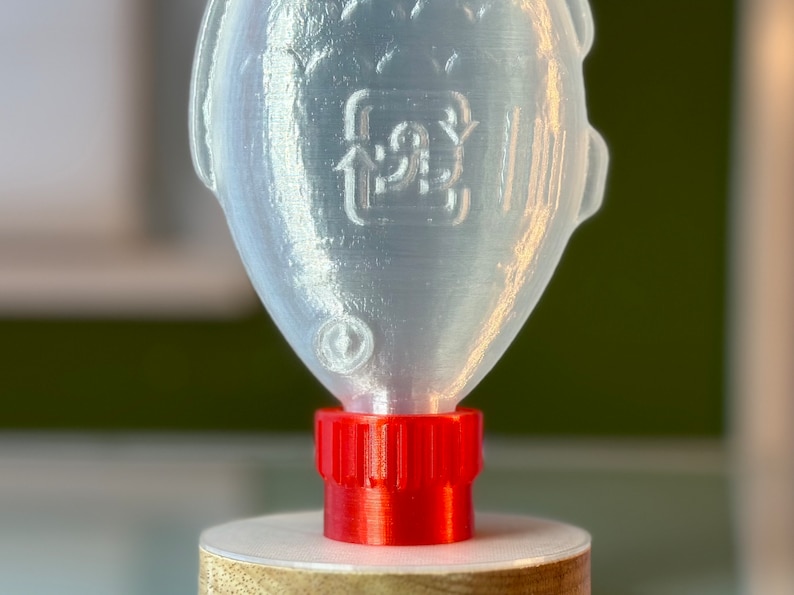May include: A clear plastic bottle shaped like a fish with a red screw-on cap. The bottle has a recycling symbol on the side.