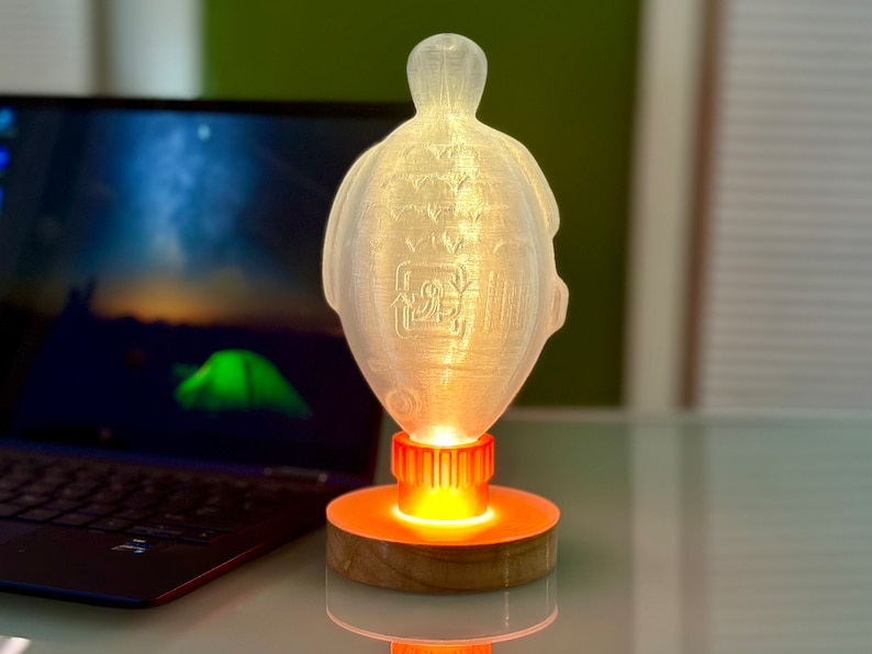 May include: A white, translucent, 3D printed lamp shaped like a turtle with an orange light base. The lamp is on a wooden base and is lit up.