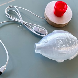 May include: A white 3D printed fish night light with a USB cord and an inline switch. The fish has a textured surface and a small hole on the side. The light is on a white surface.