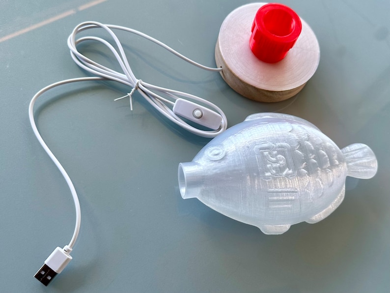 May include: A white 3D printed fish night light with a USB cord and an inline switch. The fish has a textured surface and a small hole on the side. The light is on a white surface.