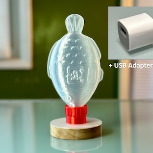 May include: A clear plastic fish-shaped bottle with a red cap, sitting on a wooden base. The bottle is designed to hold liquid and has a recycling symbol on the side. A white USB adapter block is shown in the upper right corner of the image.