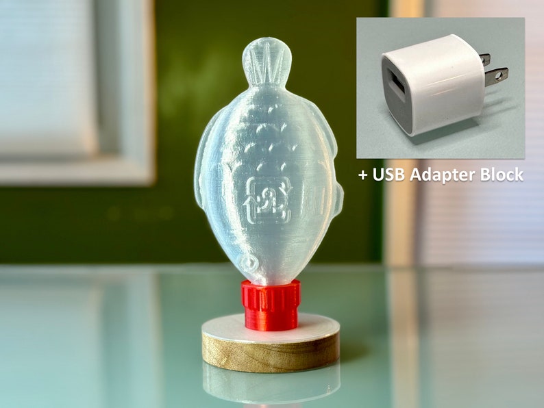 May include: A clear plastic fish-shaped bottle with a red cap, sitting on a wooden base. The bottle is designed to hold liquid and has a recycling symbol on the side. A white USB adapter block is shown in the upper right corner of the image.
