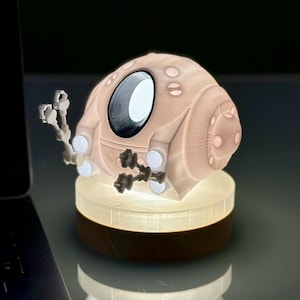 May include: A white 3D printed lamp shaped like a futuristic submarine with a round window and small robotic arms. The lamp is lit and casting a warm glow.