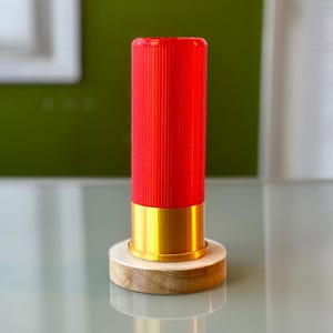 May include: A red, cylindrical object with vertical ridges, a gold-colored base, and a wooden stand. The object is standing on a reflective surface, with a green and white background.