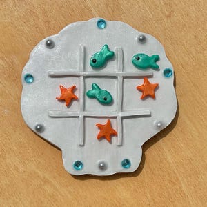 May include: A white seashell shaped tic-tac-toe game with orange starfish and turquoise fish pieces.