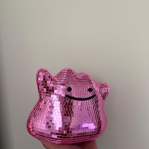 May include: A pink disco ball shaped like a blob character with a smooth, curved top and two side protrusions. The surface is covered in small, square, reflective mirror tiles. It features a simple black horizontal line for a mouth and two black dots for eyes.
