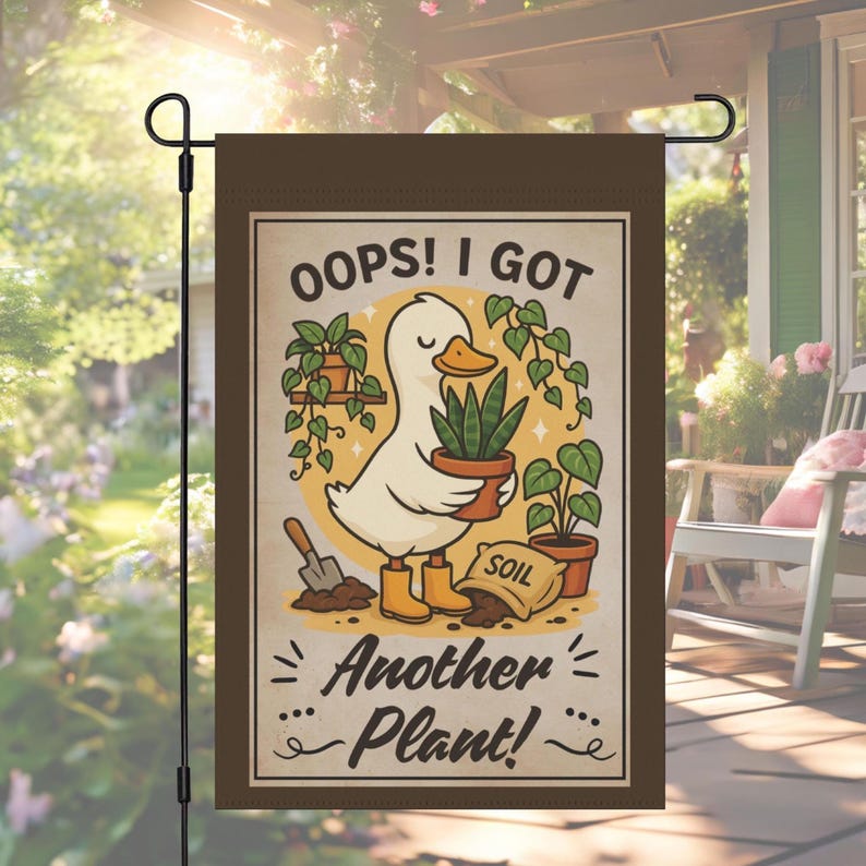 Op de afbeelding: Bruine tuin vlag met een illustratie van een eend die een potplant vasthoudt. De tekst luidt "Oops! I Got Another Plant!" in een speels lettertype. Andere planten, een schop en een zak aarde zijn ook afgebeeld.