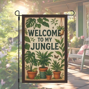 以下が含まれることがあります： 茶色の縁取りと緑色の「WELCOME TO MY JUNGLE」の文字が入った長方形のガーデンフラッグ。さまざまな鉢植えの植物、葉、カエルのイラストが特徴です。黒い金属製のポールが旗を支えています。