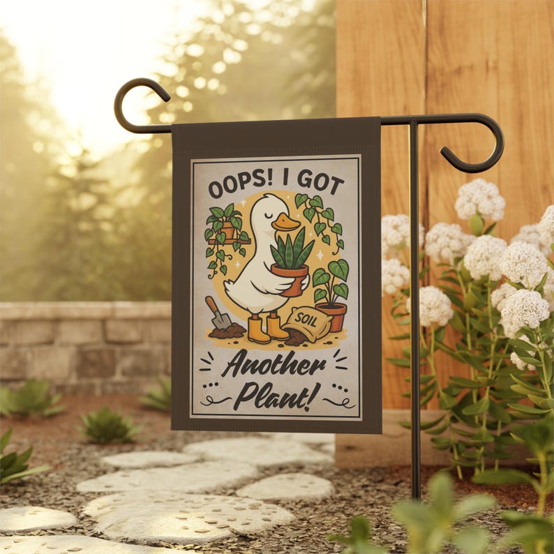 Op de afbeelding: Een tuinvaandel met een grillig ontwerp. De vlag toont een cartoon eend met een potplant, omringd door andere planten en de tekst "OOPS! I GOT Another Plant!". De vlag is gemonteerd op een zwarte metalen standaard.
