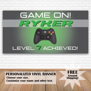 This is a video game themed vinyl birthday banner. The background is charcoal grey and lime green. There is a game controller in the middle with the text Game On Level Achieved. The name and age text is personalized.