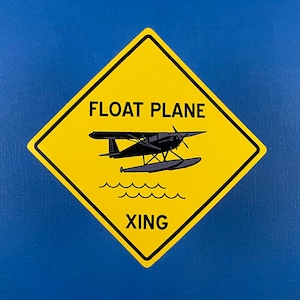 Può includere: Segnale stradale a forma di diamante giallo con bordo nero, con la scritta "FLOAT PLANE XING". Presenta un'illustrazione nera di un idrovolante sopra linee ondulate, su sfondo blu.