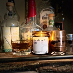 May include: A lit candle in a glass jar labeled "Bourbon & Oak" sits next to a shot of amber liquid and a copper-colored barrel-shaped candle holder. The background features various liquor bottles.