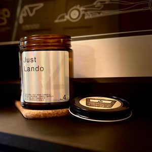 May include: A brown glass jar candle with a black lid, labeled "Just Lando." The label also indicates it's a natural soy wax candle, hand-poured in small batches, and contains 4 oz. The lid is off, and the candle sits on a cork coaster.