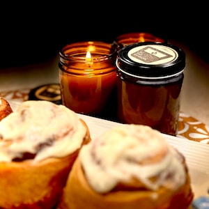 May include: A lit amber-colored candle in a glass jar, next to a closed jar with a black lid and label. Cinnamon rolls with white frosting are in the foreground. The candle flame glows brightly, creating a warm ambiance.