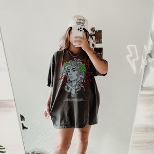 May include: A woman wearing a dark gray oversized t-shirt with a graphic print of a woman's face with snakes and the word "MEDUSA" on it. She is also wearing a white baseball cap with the text "Cowboy" on it and white sneakers.