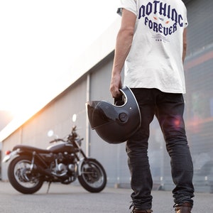 May include: A person wearing a white t-shirt with the text "NOTHING LAST FOREVER" in blue and orange lettering, black jeans, and a black baseball cap. The person is holding a black motorcycle helmet and standing in front of a black motorcycle.