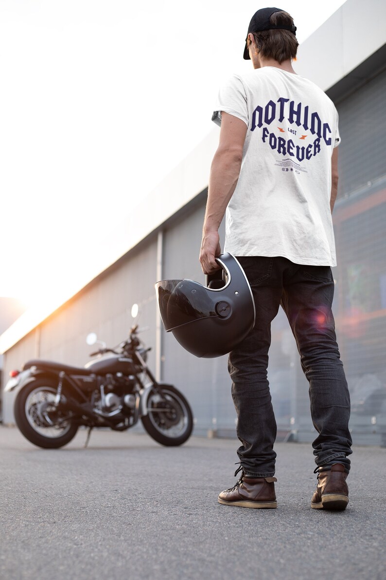 May include: A person wearing a white t-shirt with the text "NOTHING LAST FOREVER" in blue and orange lettering, black jeans, and a black baseball cap. The person is holding a black motorcycle helmet and standing in front of a black motorcycle.