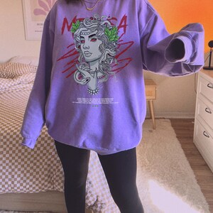 May include: Purple sweatshirt with a graphic print of a woman with snakes for hair. The graphic is in red, green, and white. The text "MEDUSA" is printed in red on the sweatshirt.