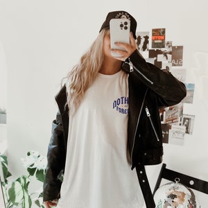 May include: A woman wearing a white t-shirt with the text "NORTH FOREVER" in blue and purple lettering, a black leather jacket, and a black baseball cap with the text "LAST GAS" in white lettering. She is holding a white iPhone in front of her face.