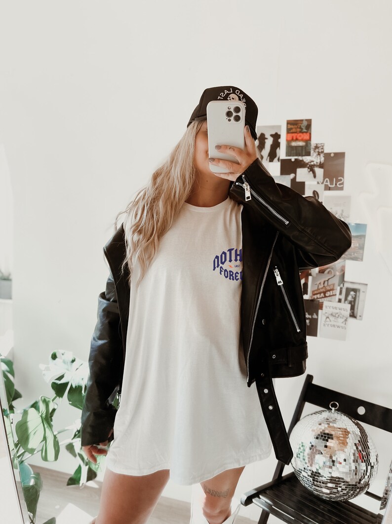 May include: A woman wearing a white t-shirt with the text "NORTH FOREVER" in blue and purple lettering, a black leather jacket, and a black baseball cap with the text "LAST GAS" in white lettering. She is holding a white iPhone in front of her face.