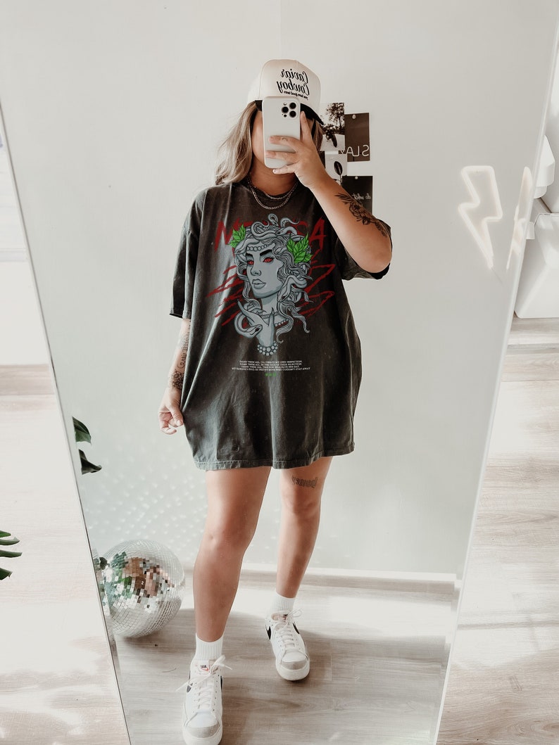 May include: A woman wearing a dark gray oversized t-shirt with a graphic print of a woman's face with snakes and the word "MEDUSA" on it. She is also wearing a white baseball cap with the text "Cowboy" on it and white sneakers.