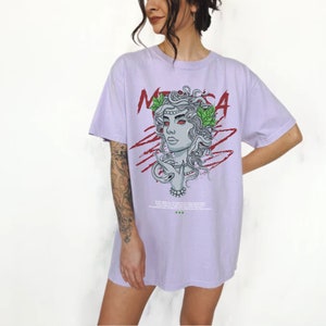 May include: A lavender t-shirt with a graphic print of Medusa, a mythical Greek figure with snakes for hair. The graphic is in black, white, red, and green. The text "MEDUSA" is printed in red on the shirt.