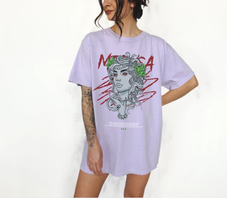 May include: A lavender t-shirt with a graphic print of Medusa, a mythical Greek figure with snakes for hair. The graphic is in black, white, red, and green. The text "MEDUSA" is printed in red on the shirt.