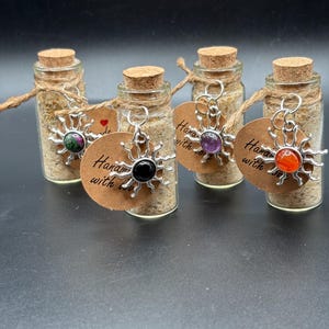 May include: Four small glass bottles with cork stoppers filled with beige sand. Each bottle features a silver-toned sun-shaped charm with a colorful gemstone center and a round brown tag printed with the text Handmade with love.