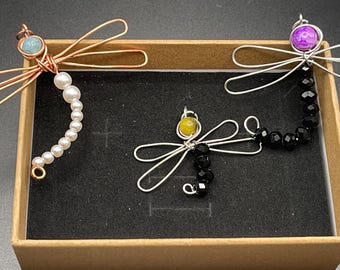 May include: Three wire dragonfly jewelry pieces with different colored beads. The first dragonfly is made of copper wire and has a light blue bead and a pearl string. The second dragonfly is made of silver wire and has a yellow bead and a black bead string. The third dragonfly is made of silver wire and has a purple bead and a black bead string.