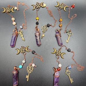 May include: A collection of amethyst crystal pendants with copper wire wrapping, decorative keys, and bronze-colored crescent moon charms. Each pendant features a chain with colorful beads. The pendants are displayed on a dark surface.