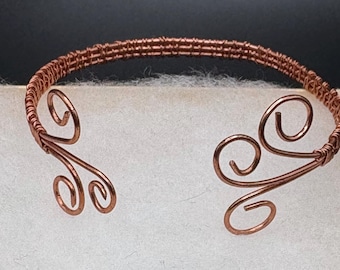 May include: Copper-colored wire cuff bracelet with an open design. The bracelet features a woven band and decorative swirls at each end. The metal has a warm, reddish-brown hue, and the overall style is delicate and artistic.