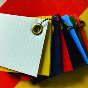 May include: A color swatch fan featuring a variety of vibrant colors, including white, yellow, red, blue, and green. Each swatch is square-shaped and attached with a metal grommet and a ball chain. The white swatch has vertical ridges.