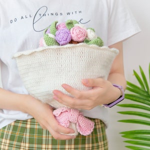 以下が含まれることがあります： ピンク、パープル、グリーンの色の毛糸で編まれた花束。花束を持っている人は、「Do all things with love」と書かれた白いシャツを着ています。