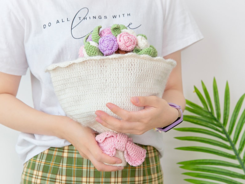 以下が含まれることがあります： ピンク、パープル、グリーンの色の毛糸で編まれた花束。花束を持っている人は、「Do all things with love」と書かれた白いシャツを着ています。