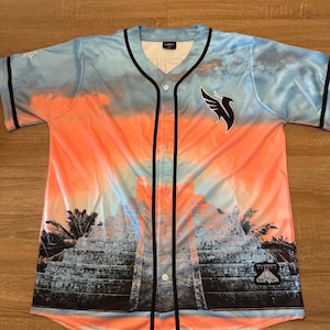 May include: Baseball jersey with a vibrant sunset design in shades of orange and blue. The jersey features a black collar, buttons, and trim. A black and white logo is on the right chest, and a graphic of a pyramid is on the front.