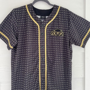 May include: Black baseball jersey with gold trim and a gold "Said the Sky" logo on the front. The jersey has a repeating pattern of gold circles on a black background.