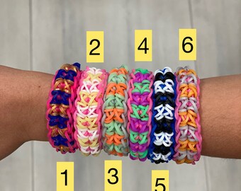 May include: Six colourful rubber band bracelets stacked on a person's wrist. The bracelets are made with different colour combinations, including pink, blue, white, orange, and black.