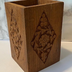 May include: A square wooden container with carved floral diamond patterns. The container is made of a dark brown wood and has a smooth, polished finish. The carved designs add a decorative element to the piece.