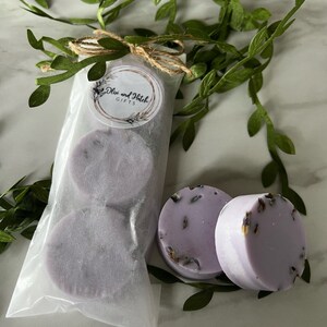 May include: Two lavender-colored bath bombs with dried lavender flowers, packaged in a white paper bag with a brown twine bow and a label that reads "Olive and Hatch Gifts".