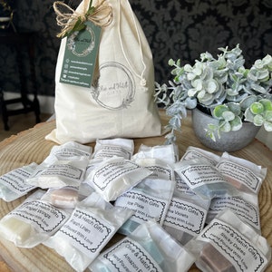 May include: A collection of small, white, paper bags filled with various coloured wax melts. Each bag has a label with the text "Olive & Hatch Gifts" and the name of the scent. The scents include "Amazon Rain", "Ice Pixie", "Fresh Linen", "Angel Wings", "Sticky Dates", "Daisy", "Lavender Escape", "Baby Powder", "Mr. Savage", and "Sea Salt & Vanilla".
