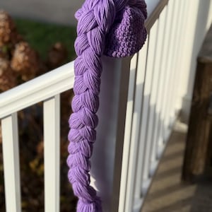 May include: A handmade, purple knit hat with a long, braided extension. The hat is resting on a white railing, with the braid hanging down. The braid is made of thick yarn and has a textured appearance. The hat and braid are a vibrant shade of purple.