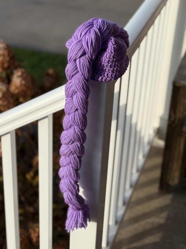 May include: A handmade, purple knit hat with a long, braided extension. The hat is resting on a white railing, with the braid hanging down. The braid is made of thick yarn and has a textured appearance. The hat and braid are a vibrant shade of purple.