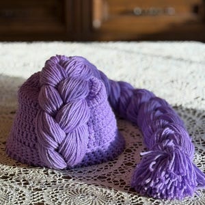 May include: A handmade, lavender-colored crochet hat with a long, braided extension. The hat features a detailed crochet stitch pattern and a thick, braided accent. The extension ends in a tassel. The hat is displayed on a white lace surface.