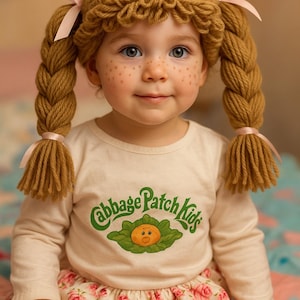 May include: A child wearing a handmade crochet wig with braided pigtails, adorned with pink ribbons. The child is wearing a long-sleeved shirt with the "Cabbage Patch Kids" logo and a floral skirt.