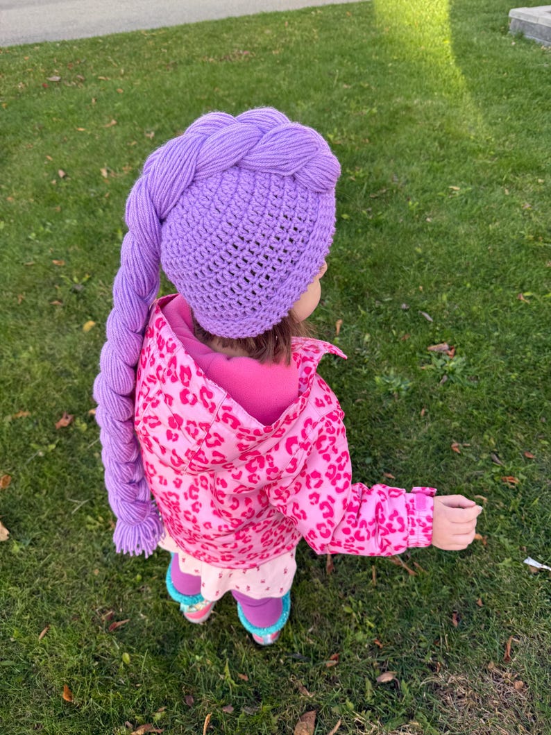 May include: A crocheted purple hat with a long braid, worn by a child. The hat is a solid lavender color, and the braid hangs down the side. The child is wearing a pink jacket with a leopard print.