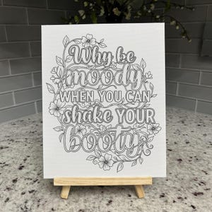 May include: A white rectangular canvas featuring black line art of floral vines surrounding the text Why be moody WHEN YOU CAN shake YOUR BOOTY. The canvas is displayed on a small, light-colored wooden easel stand for tabletop presentation.