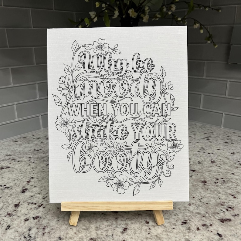 May include: A white rectangular canvas featuring black line art of floral vines surrounding the text Why be moody WHEN YOU CAN shake YOUR BOOTY. The canvas is displayed on a small, light-colored wooden easel stand for tabletop presentation.