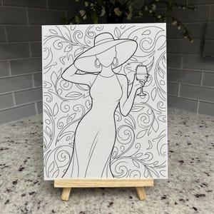 May include: A black and white line art drawing on a rectangular canvas resting on a wooden easel. The illustration depicts a figure wearing a wide-brimmed hat and a dress, holding a wine glass, set against a background of swirling floral patterns.