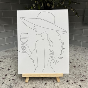 May include: A white canvas featuring a black line art illustration of a person with long wavy hair wearing a wide-brimmed sun hat and holding a stemmed wine glass. The artwork is displayed on a small wooden easel stand, showcasing a simple profile.